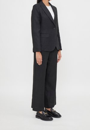 Mujer de pie vistiendo un blazer negro a medida, pantalones negros de pierna ancha, camiseta blanca y mocasines de cuero negro con hebillas doradas.