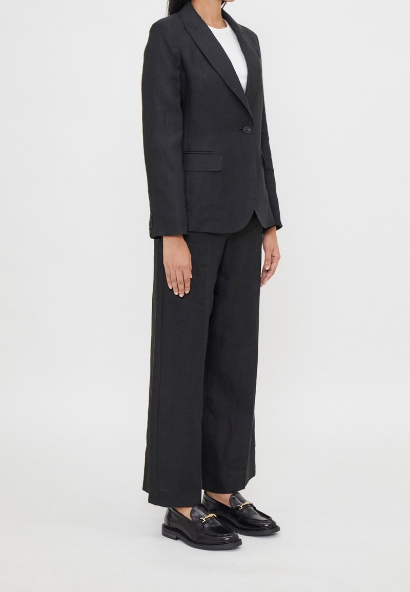 Femme debout vêtue d'un blazer noir sur mesure, d’un pantalon noir à larges jambes, d'un haut blanc et de mocassins en cuir noir avec des boucles dorées.