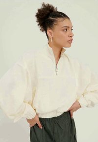 Mladá žena s kučeravými vlasmi v vysokom drdole, oblečená v krémovom zipsovom mikinovom svetri a tmavých nohaviciach s vysokým pásom, pózujúca s rukami na bokoch, hľadiaca doprava.