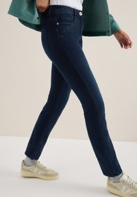 Persoon die loopt in donkerblauwe slim-fit jeans, beige sneakers met grijze sokken, een wit shirt en een gedeeltelijk zichtbaar groene jas.