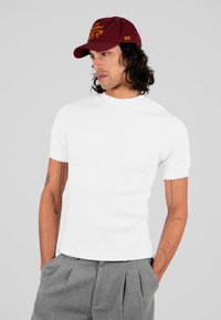 Biała krótka koszulka z ribstopem i wysokim kołnierzem, połączona z szarymi spodniami z plisami. Bordowa czapka ozdobiona haftowanym tekstem i akcentami.