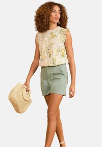 Blusa floreale con bordi in pizzo, beige con fiori gialli e verdi; abbinata a pantaloni corti verde chiaro. Tiene una borsa di paglia intrecciata.