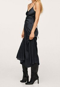 Mustassa satiinimekossa on poimutettu yläosa, säädettävät olkaimet ja virtaava epäsymmetrinen helma, joka on yhdistetty polven yli ulottuviin mustiin korkeaintyöläisiin saappaisiin.