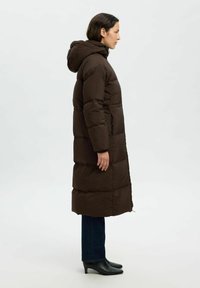 Abrigo acolchado marrón con capucha, que presenta un diseño largo y quilted. Combinado con jeans oscuros y botas de tacón negras. Textura suave.