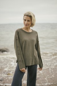Olive green knit sweater with button details on sleeves, round neckline, and relaxed fit. Paired with dark denim pants. Beach background.