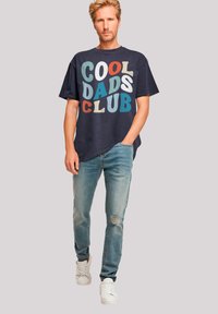 Navy cotton t-shirt featuring bold, multicolored "COOL DADS CLUB" text, paired with faded blue jeans and white sneakers.