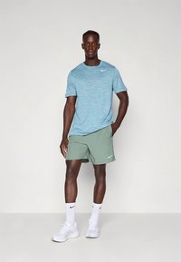 Conjunto desportivo masculino composto por uma t-shirt de manga curta azul clara mesclada e calções verdes, ambos com logotipos da Nike, combinados com ténis brancos.