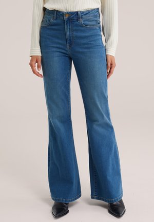 Jean bleu taille haute flare porté avec une chemise blanche à manches longues et des chaussures noires à bout pointu, visible de la taille jusqu'aux pieds.
