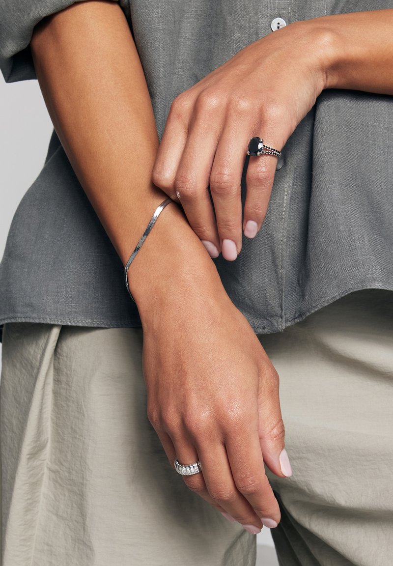 Srebrna bransoletka na jednym nadgarstku, czarny pierścionek z centralnym kamieniem i srebrną obrączką na drugim. Ręce na szarej tkaninie bluzki i jasnych spodniach.