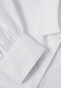 Chemise blanche ornée d'une broderie cachemire texturée sur le poignet, avec une finition lisse et un col structuré, présentant un design classique.