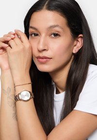 Rose gold watch with a round silver face and metal band, worn alongside a minimalistic bracelet, on a wrist with floral tattoo.
