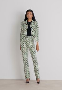 Grüner Anzug mit Muster, bestehend aus einer taillierten Jacke und passenden Hosen. Das Design zeigt geometrische Formen; getragen mit einem schwarzen Oberteil und schwarzen Absätzen.