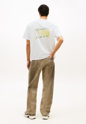 Man draagt een wit T-shirt met grafische opdruk en de tekst "Tommy Jeans" op de achterkant, bruine losse jeans en sneakers, staat met één hand in de zak.