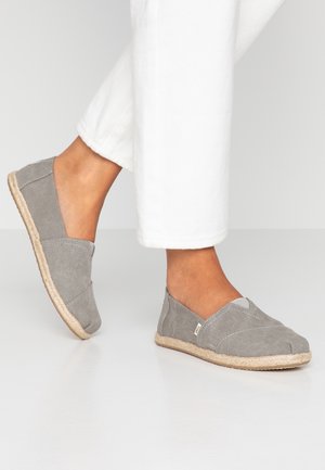 Chaussures à enfiler en toile grise avec une semelle en corde de jute, présentant un accent avant en forme de V et un tissu texturé, portées avec un pantalon blanc.