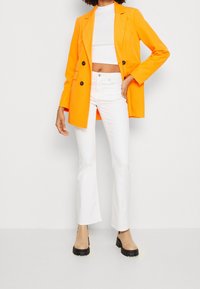 Orange dubbelknäppt kavaj, kort vit topp, vita utsvängda jeans och beige klackade ankelskor. Släta texturer, rena linjer.