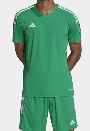 Man met een groen Adidas sportshirt en korte broek met witte strepen, staand met handen achter zijn rug op een lichte achtergrond.
