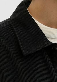 Gros plan sur une personne portant une veste en velours côtelé noir par-dessus un pull à côtes blanches, montrant les détails du col et de l'encolure.