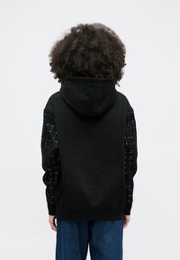 Άτομο με σγουρά μαλλιά, που φοράει μαύρο φούτερ με υφασμάτινα μαύρα μανίκια και άνετο μπλε τζιν, στέκεται με την πλάτη προς τα εμπρός σε απλό φόντο.