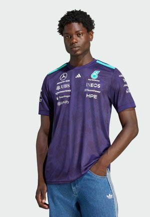 Ung mand med kort krøllet hår bærer en lilla Mercedes-AMG Petronas Formel 1 holdtrøje og blå Adidas-jeans med den ene hånd i lommen.