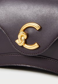 Sac à main en cuir violet foncé avec un fermoir logo doré et des accents cousus, présentant un design ondulé élégant et une texture lisse.