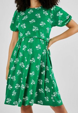Kobieta ubrana w zieloną sukienkę z białym kwiatowym wzorem i krótkimi bufkami na rękawach, z ręką opartą na biodrze na jednolitym tle.