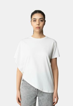 Donna con capelli scuri raccolti indossa una maglia bianca asimmetrica a maniche corte e jeans grigio chiaro su uno sfondo chiaro e uniforme.