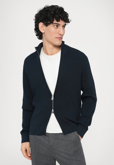 Μπλε ριγωτό πουλόβερ με φερμουάρ και όρθιο γιακά, σε συνδυασμό με λευκό μπλουζάκι με στρογγυλή λαιμόκοψη. Το μοντέλο φορά γκρι παντελόνια. Ορατή ασημί λεπτομέρεια φερμουάρ.