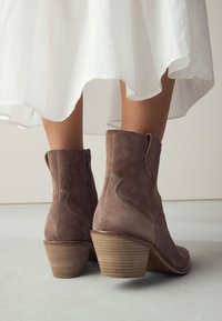 Brune ankelstøvler i ruskind med en tekstureret stablet hæl, lynlås i siden og dekorative sømme, kombineret med et flydende hvidt skørt.