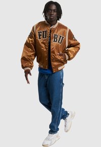 Bronze satin varsityjakke med sort broderet skrift, stribede ribkunder, parret med blå denimbukser og hvide sneakers.