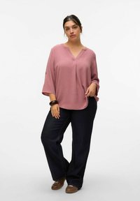 Blusa rosa texturizzata con scollatura a V e maniche ampie, abbinata a pantaloni in denim scuro e scarpe marroni. Design semplice e casual.