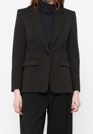 Femme portant un blazer noir cintré avec une fermeture à un bouton, des poches à rabat, et un pantalon noir à jambes larges sur fond blanc.