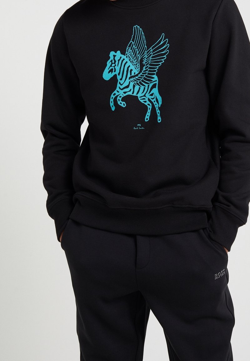 Sweat-shirt noir avec un motif zèbre ailé bleu sur la poitrine, porté avec un pantalon noir, mains dans les poches.