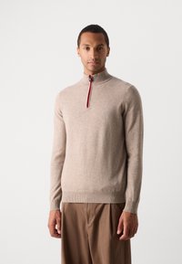 Tan, zip-up sweater made of soft fabric with a high collar. Features a red and blue accent along the zipper and ribbed cuffs.