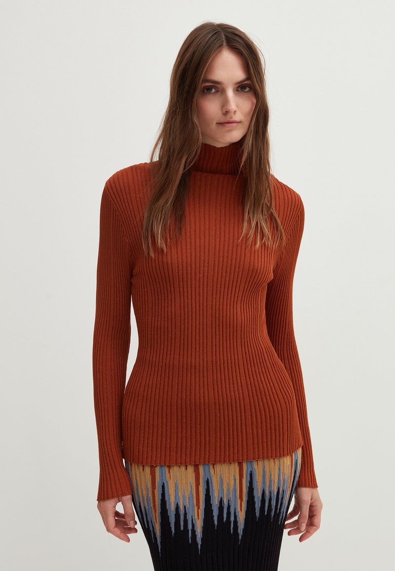 Rippstrukturierter Rollkragenpullover in gebranntem Orange, mit einer taillierten Silhouette und langen Ärmeln, kombiniert mit einem Rock im mehrfarbigen Zickzackmuster.