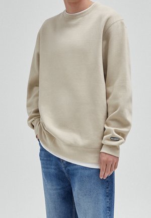 Osoba w beżowej bluzie crewneck z naszywką na rękawie i niebieskich dżinsach, stojąca z jedną ręką w kieszeni.