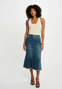 Gonna midi in denim con orlo svasato, caratterizzata da cuciture verticali e un bordo grezzo. Abbinata a una canotta a righe chiara e sandali con tacco marroni.