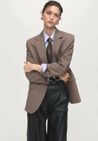 Bruine houndstooth blazer over een blauwe shirt en donkere stropdas, gecombineerd met zwarte geplooide neplederen broek. Grote gouden oorbellen aanwezig.