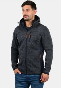Chaqueta de forro polar gris oscuro para hombre con capucha, paneles en contraste en color marino, bolsillos con cremallera y detalles en cuero marrón en las cremalleras.