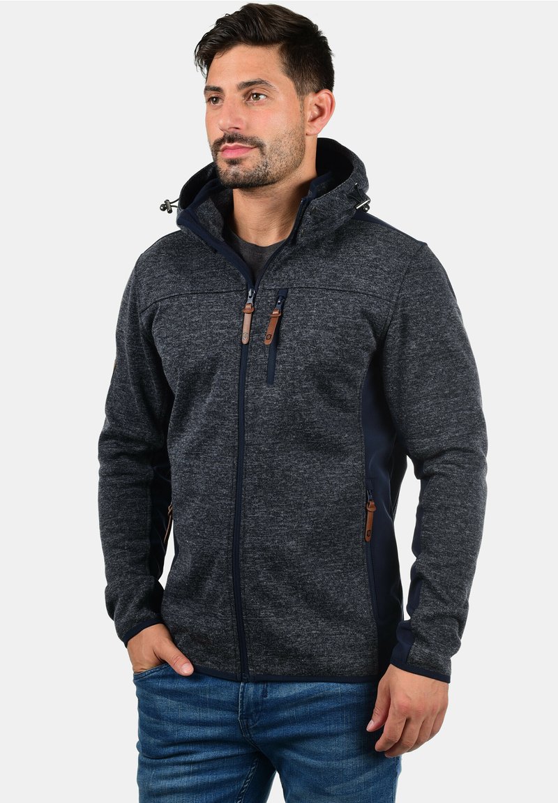 Chaqueta de forro polar gris oscuro para hombre con capucha, paneles en contraste en color marino, bolsillos con cremallera y detalles en cuero marrón en las cremalleras.