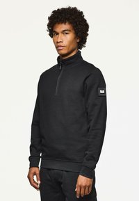 Sweatshirt negru cu fermoar, fabricat dintr-un amestec de bumbac. Prezintă un guler înalt, manșete ribbate și un petic pe brațul stâng. Textură netedă.