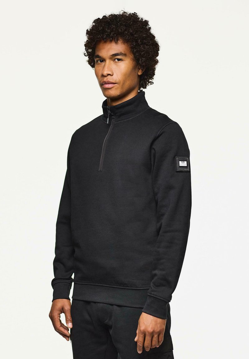 Sweatshirt negru cu fermoar, fabricat dintr-un amestec de bumbac. Prezintă un guler înalt, manșete ribbate și un petic pe brațul stâng. Textură netedă.