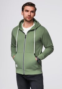 Grøn zip-up hoodie med en snørehætte, frontlommer og hvide accenter. Båret over en hvid skjorte med mørke jeans.