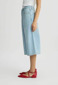 Culottes din denim de un albastru deschis, cu croială largă și scurtă, asortate cu balerini din piele roșie, cu detaliu cu fundă și bretele la glezne.