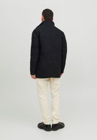Abrigo negro de corte recto con un cuello alto, de tejido texturizado, acompañado de pantalones color crema y zapatos negros. Fondo liso.