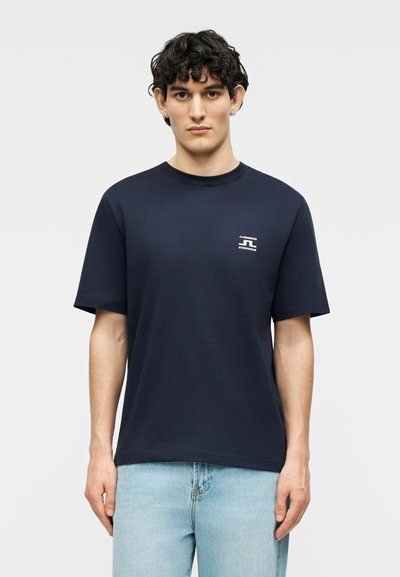 Νεαρός με σγουρά μαλλιά που φοράει μπλε μπλουζάκι με το λογότυπο J.Lindeberg Stockholm και ανοιχτόχρωμο τζιν, στέκεται μπροστά σε ουδέτερο φόντο.