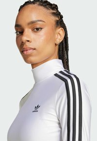Valkoinen pitkähihaista paitaa, jossa on korkea kaulus, kolme mustaa raitaa olkapäillä ja pieni musta adidas-logo rinnassa.
