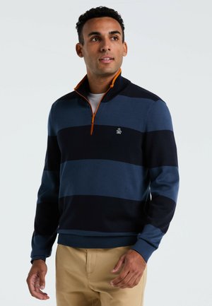 Mann trägt einen blau-schwarz gestreiften Viertelreißverschluss-Pullover mit orangefarbenem Reißverschluss und beige Hosen und schaut leicht zur Seite.