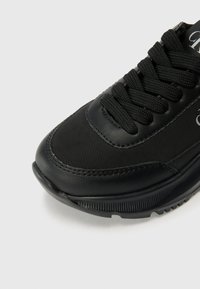 Baskets noires avec un dessus en cuir lisse et textile, bout rond, lacets plats noirs et une semelle en caoutchouc texturée. Détail de marque subtil.