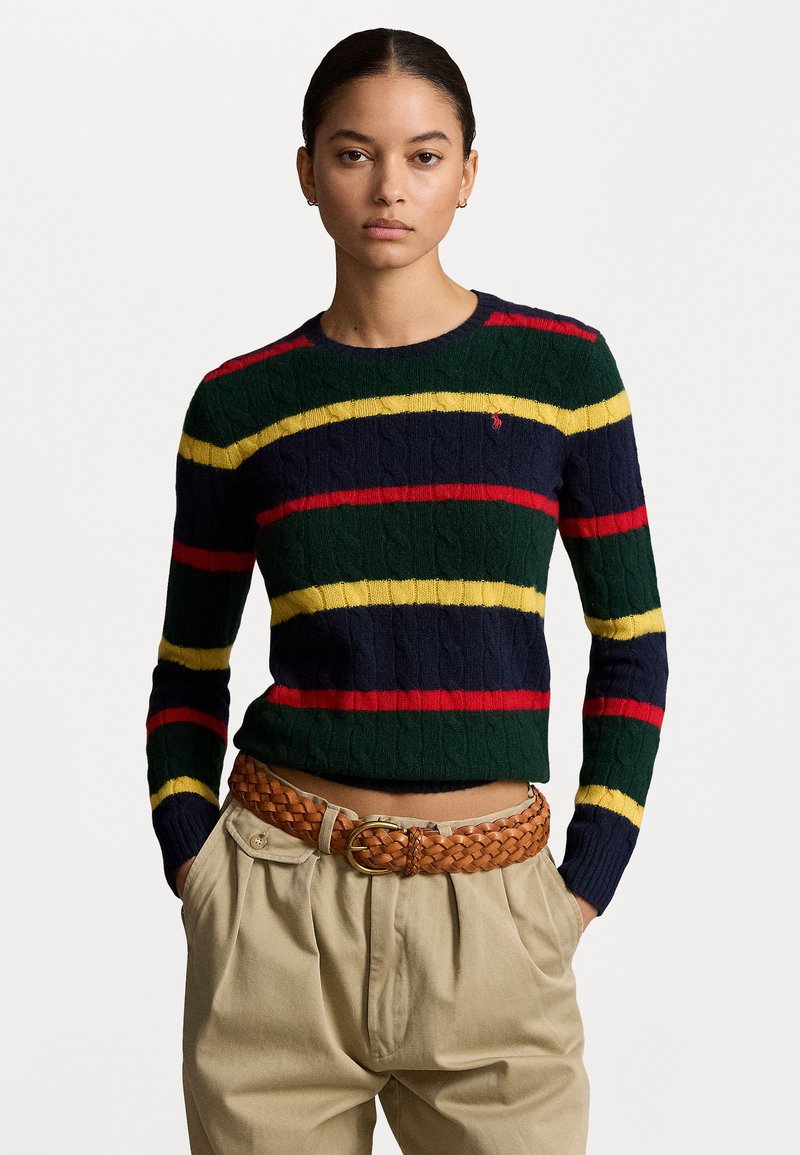 Pull en tricot avec des rayures bleu marine, vert, rouge et jaune. Présente une texture en tricot torsadé et un col rond. Associé à un pantalon beige taille haute et une ceinture tressée.