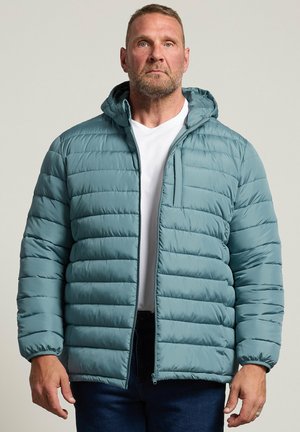 Homme portant une veste matelassée bleu clair avec capuche, t-shirt blanc et pantalon bleu foncé, debout devant un fond uni.